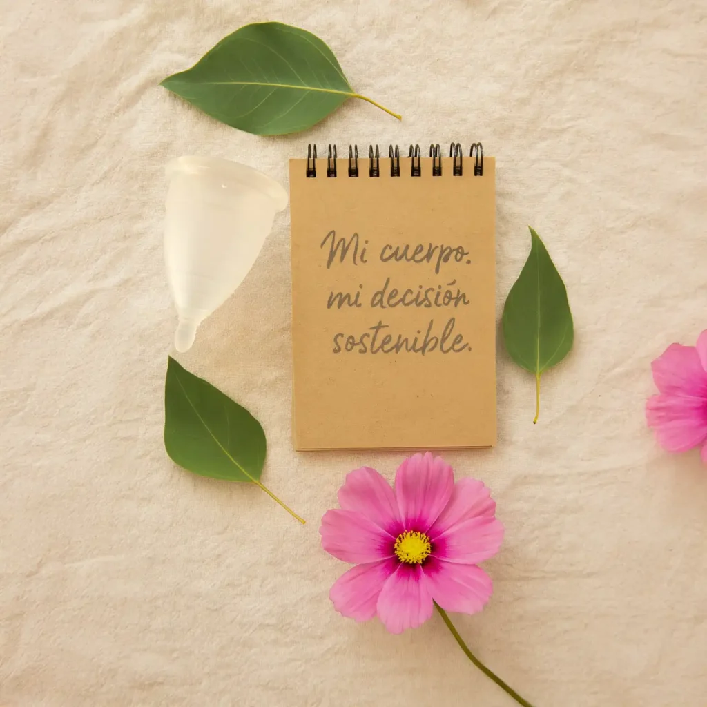 Flatlay con copa Angelcup (color cristal, lila o rosa) sobre fondo de tela orgánica o lino natural, acompañada de hojas verdes, una flor y una libreta con la frase “mi cuerpo, mi decisión sostenible