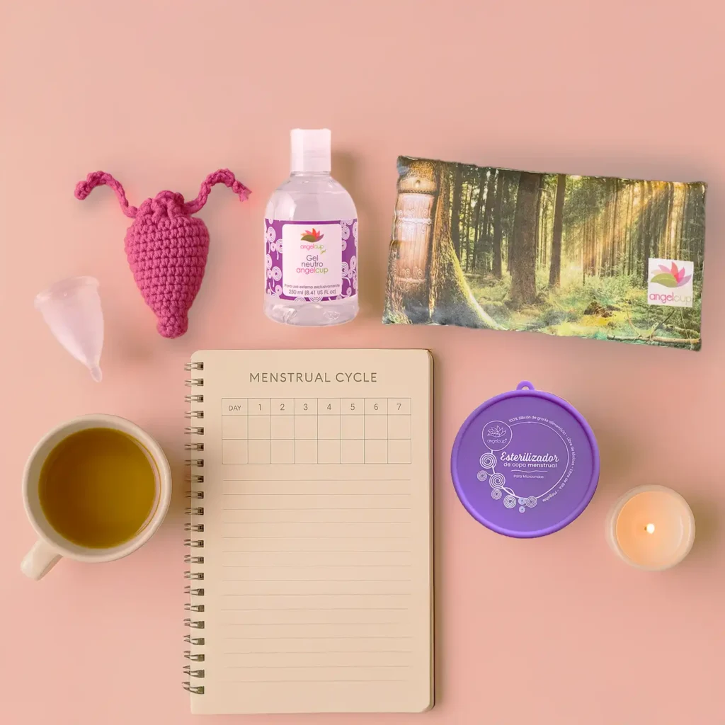 Foto sobre fondo blanco suave, con los productos Angelcup distribuidos armoniosamente: copa con su bolsa, gel neutro, cojín anticólicos doblado con flor encima, vaso esterilizador abierto. Acompañado de una libreta de registro menstrual, una vela encendida y una taza de té.