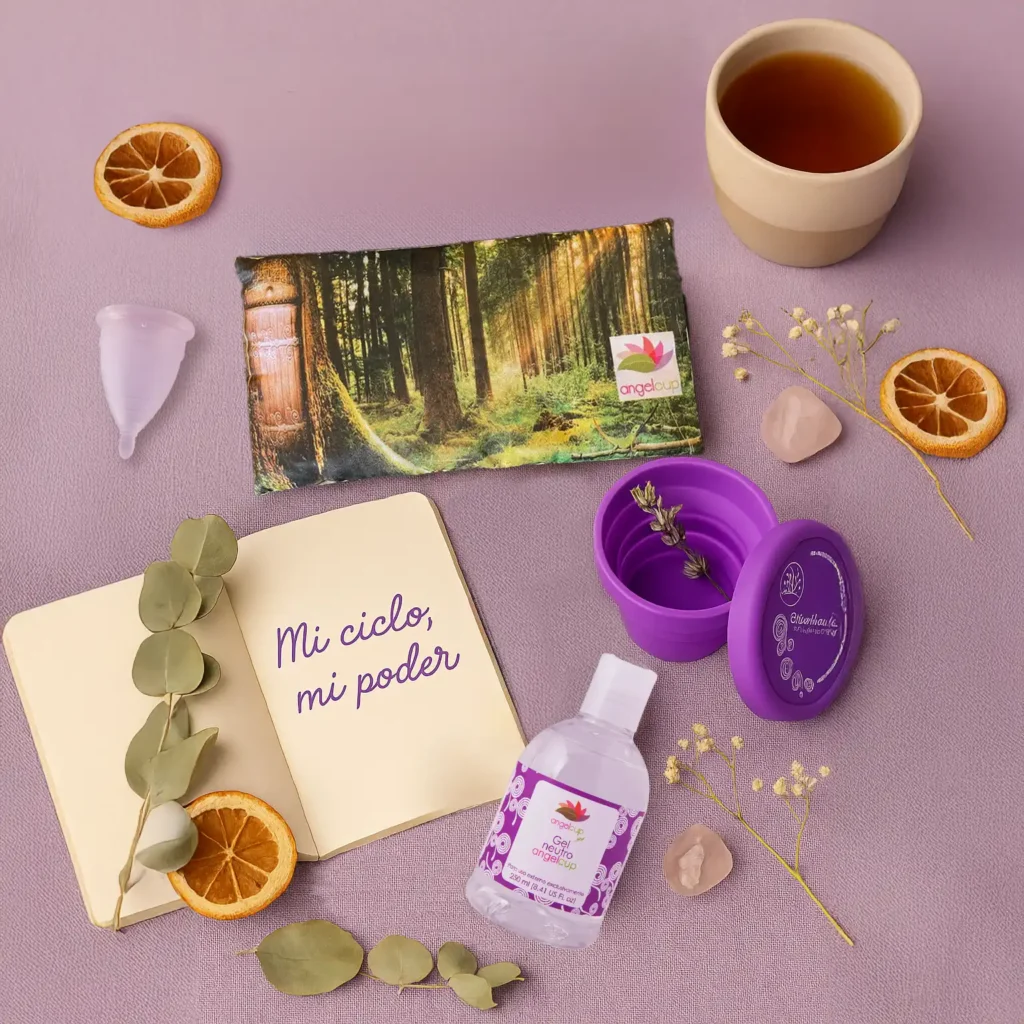 Kit de cuidado menstrual Angelcup sobre fondo lila: incluye copa menstrual, vaso esterilizador morado, gel neutro, cojín térmico con diseño de bosque, una libreta con la frase 'Mi ciclo, mi poder', y elementos naturales como té, eucalipto, flores secas, rodajas de naranja y piedras rosas. La imagen transmite bienestar, conexión cíclica y autocuidado consciente