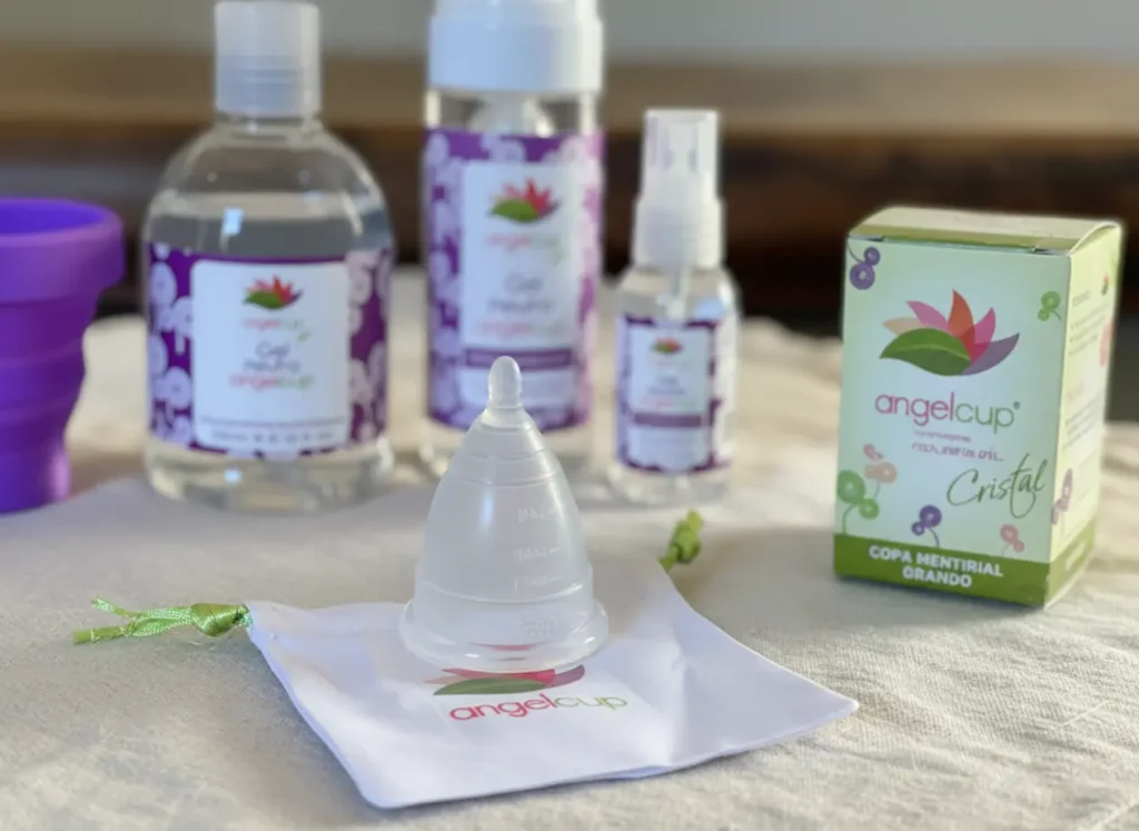 Imagen de productos Angelcup sobre una superficie clara: copa menstrual cristal sobre su bolsita de tela blanca con logotipo, caja ecológica verde y geles neutros con etiquetas moradas. Una composición que transmite limpieza, confianza y calidad mexicana.