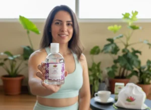 Mujer en ropa deportiva sosteniendo un frasco de Gel Neutro Angelcup hacia la cámara, con una expresión amable y segura. Detrás se aprecian plantas y una mesa con una taza, una bolsa con el logo de Angelcup y una caja del producto, creando un ambiente natural y acogedor que refleja emprendimiento consciente y bienestar.