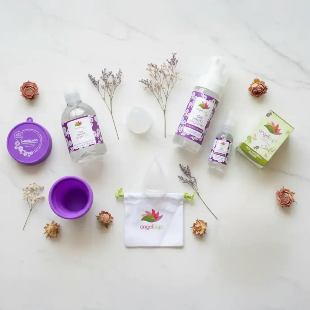 Arreglo flat lay de productos Angelcup sobre fondo blanco: copa menstrual, gel neutro, vaso esterilizador, spray limpiador, caja de presentación y bolsa de algodón con el logo de la marca. Decorado con flores secas que evocan limpieza, naturaleza y bienestar.