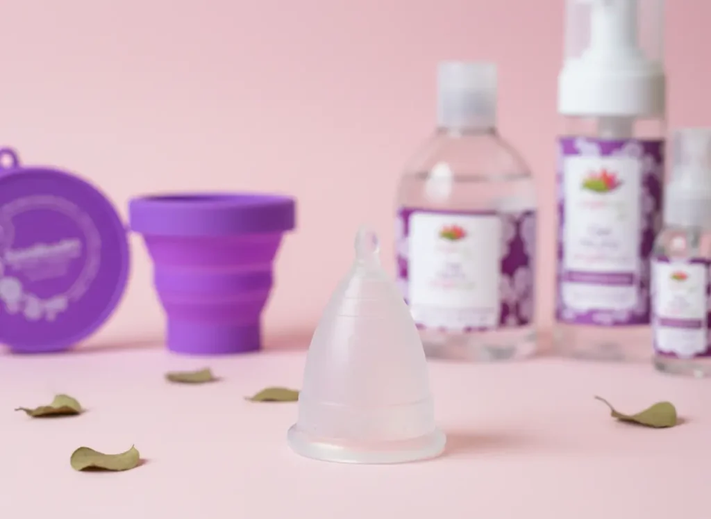 Copa menstrual Angelcup cristal en primer plano junto a esterilizador plegable morado y gel íntimo, sobre fondo rosa suave, mostrando un kit de autocuidado femenino ecológico.