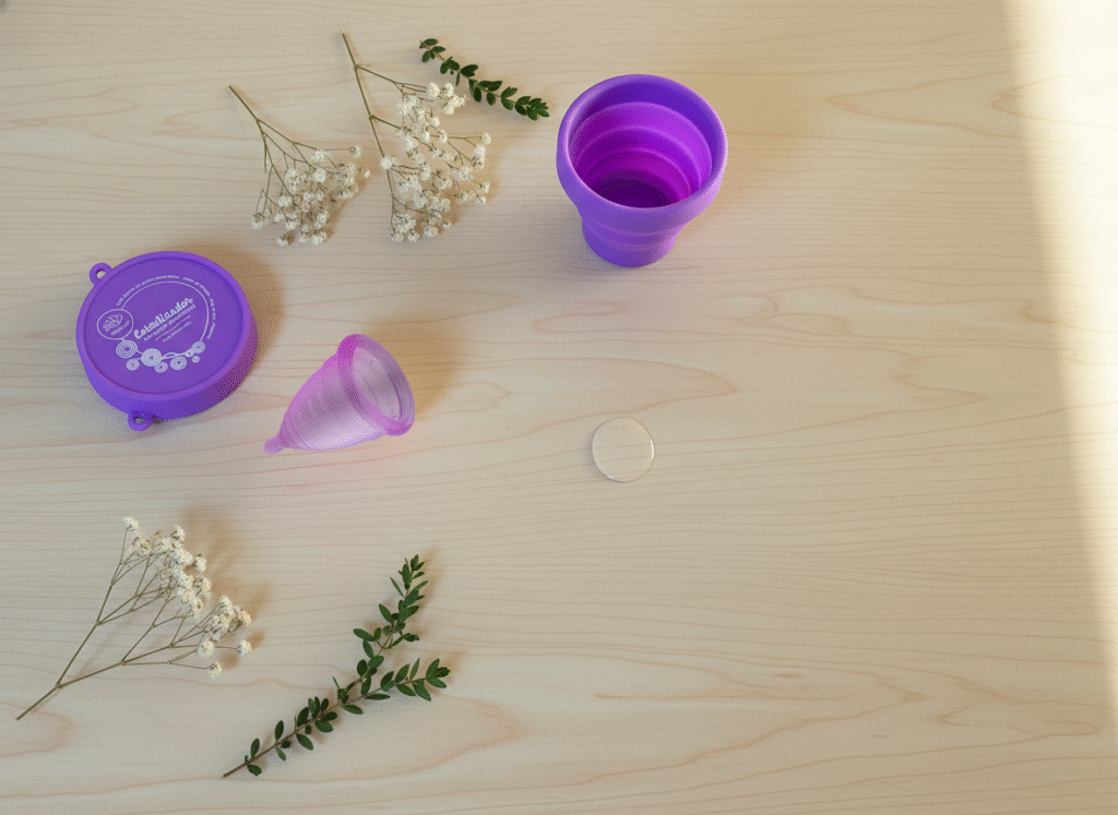 Copa menstrual Angelcup lila con esterilizador morado sobre mesa de madera clara, acompañados de flores secas y luz natural, representando autocuidado menstrual, higiene íntima y bienestar femenino sostenible.
